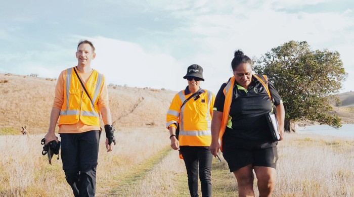 Three people, two in high vis wear, walk side by side across a rural grassland.