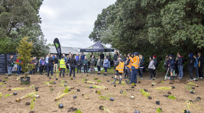 Plants are laid out in the foreground ready to plant. Behind stands a group of volunteers listening to organisers.